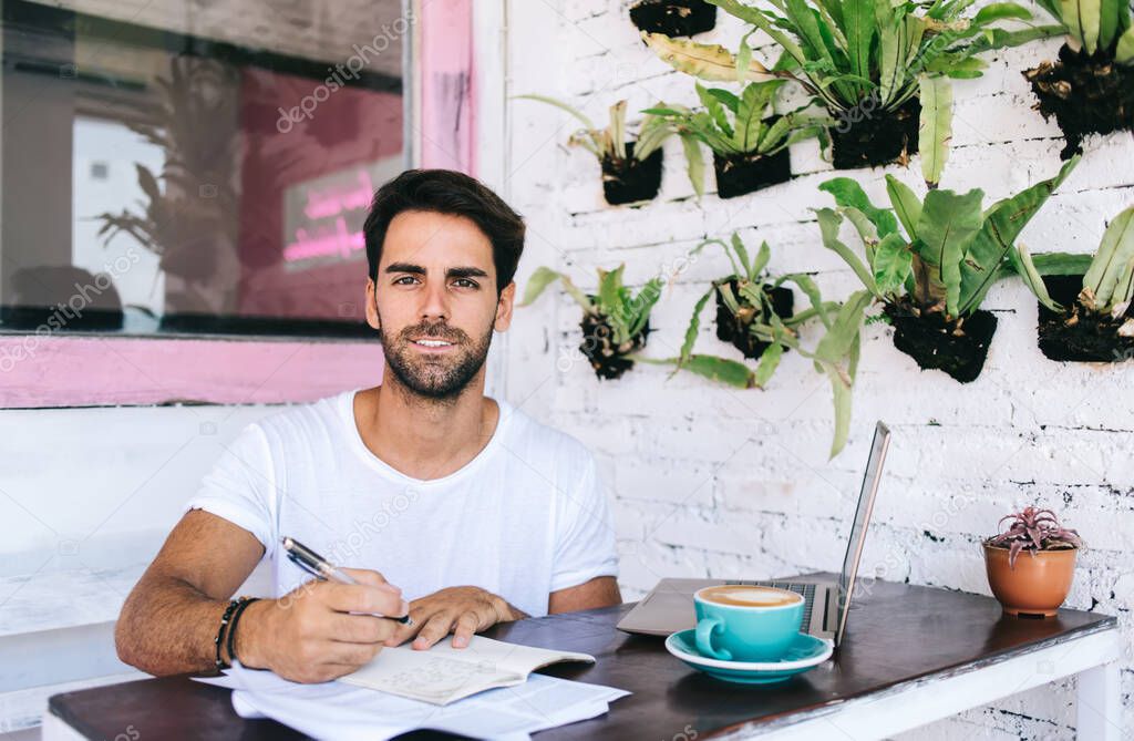 Portrait of cheerful caucasian male blogger making notes of article for share in social networks working in cafeteria, smiling creative man deschner drawing sketches in notebook on remote job