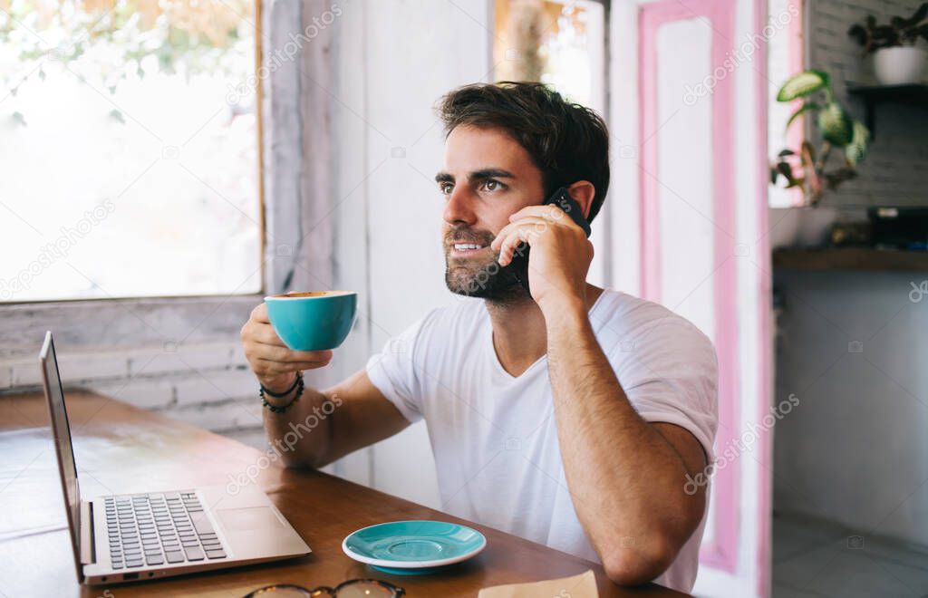 Smiling caucasian male holding coffee cup on break talking on mobile phone and working remotely, positive handsome hipster guy enjoying spending time in cafe interior making smartphone conversation