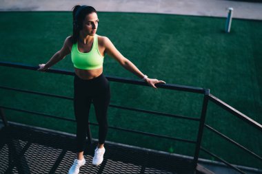 Spor kıyafetlerinde koşan düşünceli bir sporcu kulaklıkla müzik dinlerken korkuluklara yaslanıp spor sahasına bakıyor.