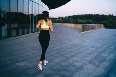 Modern binanın yakınında koşan ve kulaklıkla müzik dinleyen modaya uygun spor giyim ve beyaz spor ayakkabılı tanınmamış zayıf kadın koşucunun arka planı.