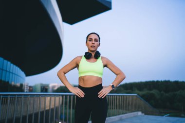 Spor kıyafetli, kulaklıklı, kameraya bakan ve ellerin belde durarak yaz sabahı sokakta çalışırken ayakta duran formda genç bir kadın.