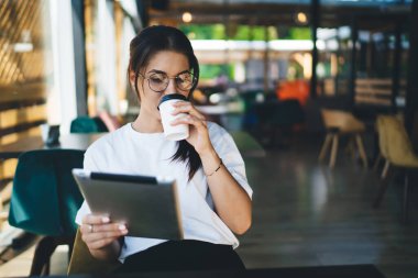 Görme düzeltmesi için gözlüklü genç kadın 4G internete bağlı tablet online kitap okuyor, sosyal ağlarda haber ve e-postaları kontrol eden hippi kız blogcu kahve içiyor.