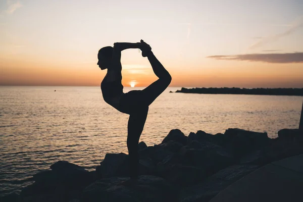 全身で見るシルエットの若い女性練習ヨガの練習海辺で海の岩場に立つ主のダンスポーズ日没時 — ストック写真
