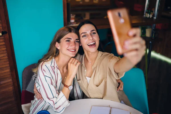スマートフォンのカメラを見ている白人の影響力のブロガーや笑顔をクリックしながら自撮り画像 幸せな女性の千年紀20代一緒にコーヒーショップで余暇の間に携帯電話の画像を作ります — ストック写真