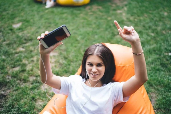 幸せな白人の若い女性は 公園で無料の時間に音楽を楽しみながら 携帯電話でプレイリストに踊り スマートフォンやイヤフォンを使用して20代のかなり肯定的な女性を笑顔オンラインラジオ — ストック写真