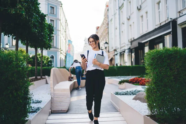 現代的な建物の近くの通りを歩いている間 バックパックブラウジングスマートフォンと保持ノートパッド付きのカジュアルな服の幸せな若い女性 — ストック写真