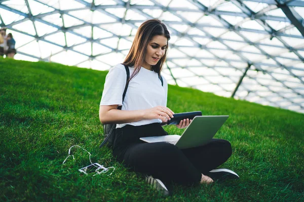 穿着休闲装的女人在几何当代建筑附近的草坪上边走边用腿远程工作边浏览笔记本电脑 — 图库照片