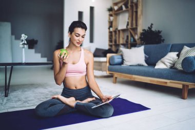 Spor kıyafetleri giymiş, yoga minderinde poz veren ve tablet kullanırken yeşil elma yiyen sakin, zayıf kadın.