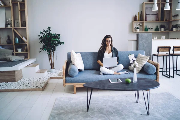 Full body of woman in casual wear using laptop and sitting with legs crossed on sofa while working on project at home