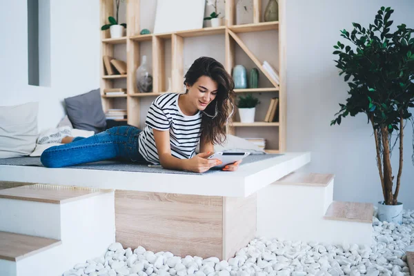 Full body of cheerful young female in casual outfit lying on floor near bookshelf and browsing tablet at modern apartment