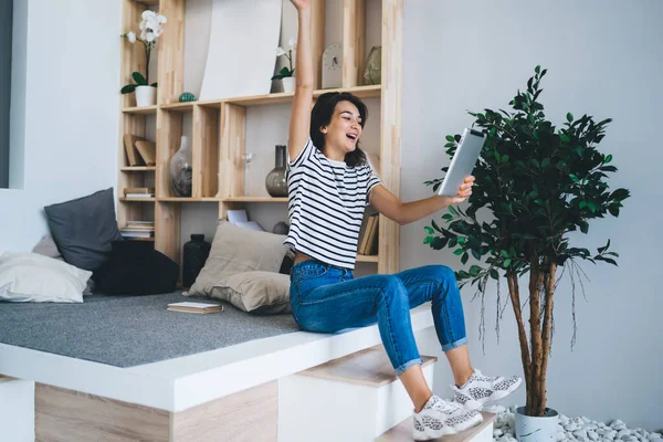 Full body of happy encouraged young female in casual outfit greeting with hand while having video conversation with friends on tablet during quarantine