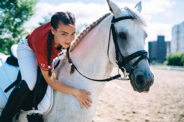 Süvari kıyafetli çekici bir kadın at sırtında oturuyor ve güneşli bir günde otlakta beyaz atın boynuna yaslanıyor.