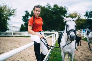 Siyah çizmeli, kırmızı gömlekli, beyaz atın yanındaki metal bariyerde oturan ve kameraya bakarken dizginleri tutan gülümseyen kadın binici.
