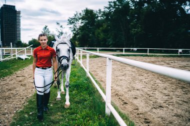Siyah çizmeli ve kırmızı gömlekli kadın jokey bariyerin yanındaki çimlerin üzerinde beyaz atla yürüyor.