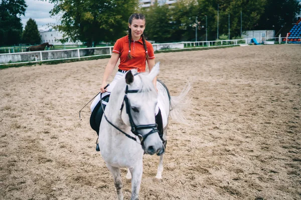 Young female equestrian in red uniform riding white horse on sandy arena on cloudy summer day during training before competition