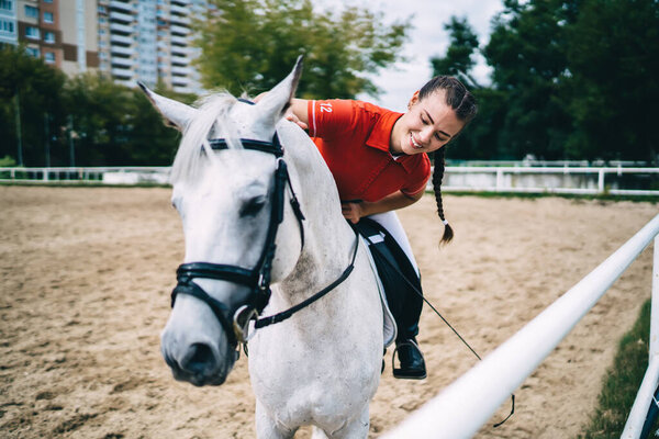 Caucasian female jockey looking at breed white stallion during horseback riding in country club with courses in paddock, cheerful woman feeling happiness during dressage with thoroughbred animal