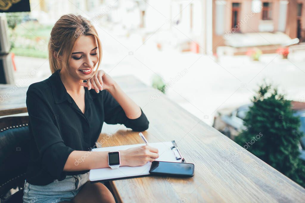 Cheerful female student with candid smile planning organisation on leisure in sidewalk cafeteria, happy Caucasian hipster girl 20 years old with papers writing project ideas enjoying good day in city