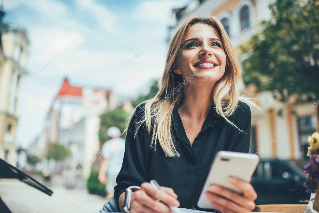 Cheerful female with cute smile on face holding cellular device for browser networking enjoying leisure in city, prosperous hipster blogger feeling carefree happiness and delight from mobility