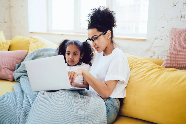 African American mother in white shirt with eyeglasses sitting on sofa with adorable daughter together under blanket and watching movie on laptop