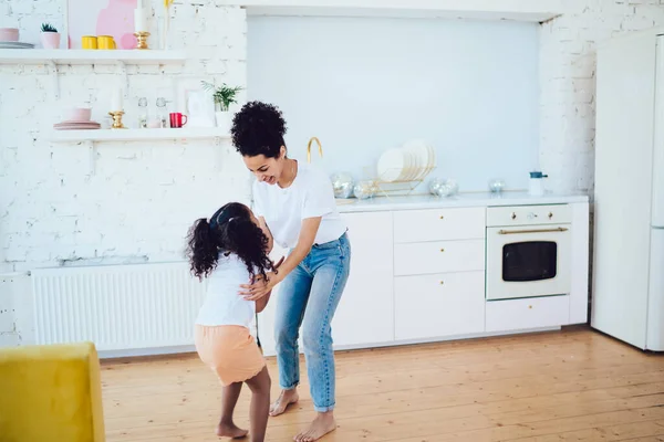 笑顔アフリカ系アメリカ人の母親でカジュアルな服ともに巻き毛ちびっ子笑って娘同時に立っています現代的なキッチンのスタジオアパート — ストック写真
