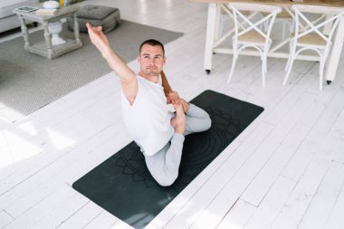 Sıradan giyinmiş sakin, beyaz bir adam Asana 'da oturmuş evde sağlıklı zihin ve bedenin tadını çıkarıyor. Yoga ve meditasyondan tatmin olan genç bir erkek halı üzerinde egzersiz yapıyor.