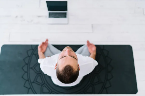Top view of male sitting on carpet with mandala enjoying meditation and recreation at home interior, caucasian man sitting in lotus pose keeping healthy lifestyle and mind relaxing in apartment