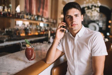 Normal giyinen genç adam, bar tezgahının yanındaki restoranda kırmızı kokteyl içerken akıllı telefondan konuşuyor ve kameraya bakıyor.