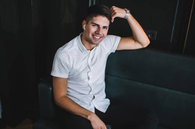 Gerçek kablosuz kulaklıklı, rahat koltukta dinlenen, başı öne eğik, hafta sonu dinlenirken kameraya bakıp gülümseyen genç ve yakışıklı bir erkek.