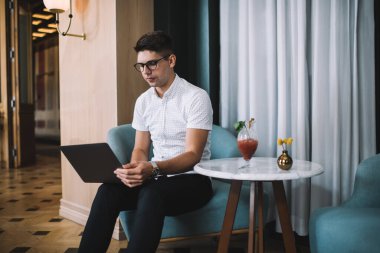 Resmi giyinmiş, gözlüklü genç bir erkek öğrenci kafede otururken dizüstü bilgisayardan bilgi arıyor.