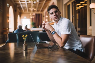 Sıradan giysiler içinde, elinde kalemle kafede dizüstü bilgisayarla çalışırken durup başka tarafa bakıyor.