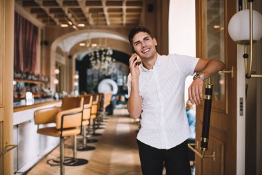 Cep telefonuyla konuşurken ayakta durup kapıya yaslanırken gülümseyerek ve yüzünü çevirerek gülen neşeli genç erkek.