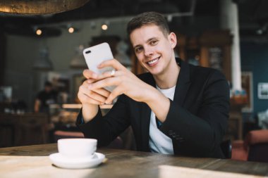 Mutlu, genç, zarif giyinmiş, gülümseyen ve kameraya bakan bir erkek kafeteryada akıllı telefon ve bir fincan sıcak içecekle masada oturuyor.