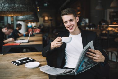 Mutlu gülümseyen, iyi giyimli, kahve içen ve gazete okuyan bir erkek. Bu arada cep telefonu ve not defteriyle karanlık, rahat bir kafede oturuyor.