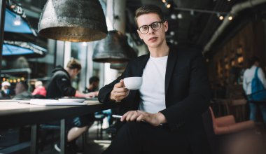 Kahve masasında oturmuş elinde kalemle kameraya bakarken beyaz tişörtlü ve siyah takım elbiseli ciddi bir genç kahve içiyor.