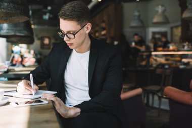 Konsantre olmuş, koyu saçlı, resmi takım elbiseli, gözlüklü, kafede otururken not defterine yazan genç bir erkek.
