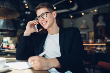 Gündüz vakti kafede not defteriyle otururken şık Google 'larda ve günlük kıyafetlerle gülümseyen erkek öğrenci ödevlerini yapıyor.
