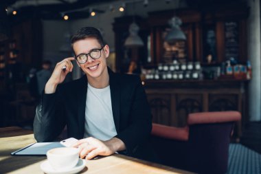 Resmi kıyafetler içinde, gözlüklü, kulaklarına dokunup kameraya bakarken cep telefonu ve kahve içen mutlu bir erkek yönetici.