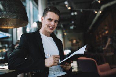 Rahat bir kafede oturup, proje üzerinde çalışırken ve kameraya bakarken günlük planlamada notlar alan akıllı, rahat bir erkek yönetici.