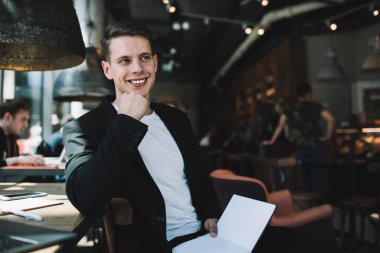 Kalabalık bir kafede oturan ve uzaktan yeni bir proje üzerinde çalışırken gülümseyen ve başka yöne bakan memnun erkek girişimci