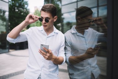 Gözlüklü, beyaz tişörtlü, gözlüklü, internette cep telefonuyla sörf yapan genç adam gün ışığında şeffaf duvara yaslanmış ve başka tarafa bakıyor.