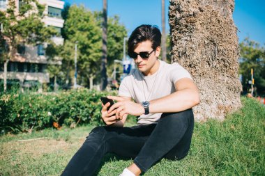Güneş gözlüklü ciddi bir beyaz adam akıllı telefonuyla haberleri okuyor. Şehir parkında boş vakitte oturuyor. Güneşli bir günde açık havada 4G kullanarak cep telefonunda yakışıklı bir erkek sözü geçen internet sitesi var.