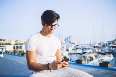 Moda gözlükleri ve günlük kıyafetleriyle deniz kenarındaki taştan sınırda oturan ve akıllı telefondan arkadaşlarıyla sohbet eden ciddi stil sahibi bir adam.