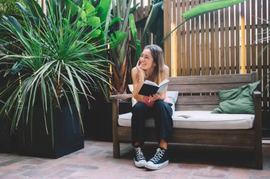 Haftasonu dinlenirken ahşap bankta oturmuş kitap okuyan, günlük giysiler içinde neşeli bir kadın vücudu.