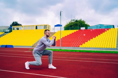 Stadyumda koşuya başlamadan önce eşofmanlı, uzun boylu, kararlı bir erkek koşucu ısınıyor ve geriye doğru hareketler yapıyor.