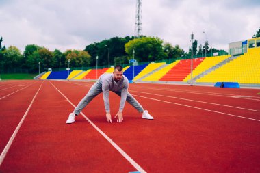 Genç kararlı erkek koşucu sabah antrenmanında mükemmel vücut şeklini koruyor, 20 'li yaşlarda aktif giyimli ciddi bir sporcu stadyumdaki kardiyo eğitiminde alt vücut kaslarını esnetiyor.
