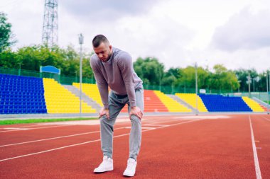 Genç beyaz erkek koşucu stadyumda ayakta duruyor. Aktif kardiyo eğitiminden sonra nefes alıyor. Egzersiz sırasında vücudunu formda tutmak ve formda kalmak için çalışan bitkin hippi.