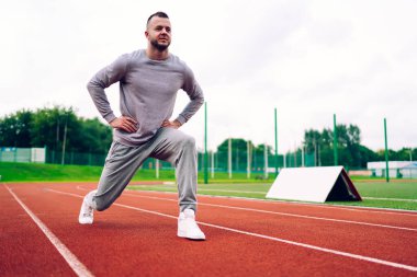 Gri spor giyim ve spor ayakkabılarıyla spor yaparken ve stadyuma bakarken esneme egzersizi yapan kararlı sporculardan oluşan bir vücut.