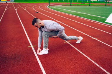 Ciddi güçlü erkek koşucu esnek vücut için bacakları çalıştırmadan önce egzersiz yapıyor, modaya uygun eşofmanlı sporcu fitness hedeflerine ve sağlıklı aktif yaşam tarzını sürdürüyor.