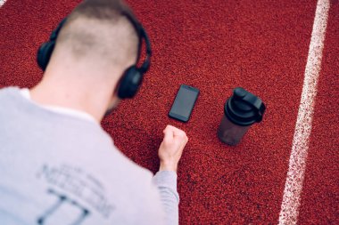 Yüksek açılı, kulaklıklı, akıllı telefon ve su şişeli genç erkek sporcu lastik iz üzerinde düşük kalas egzersizi yapıyor.