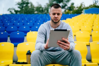 Boş stadyumun koltuğunda dinlenen ve tablet üzerinde çalışan sıradan giysili, düşünceli genç bir adam.